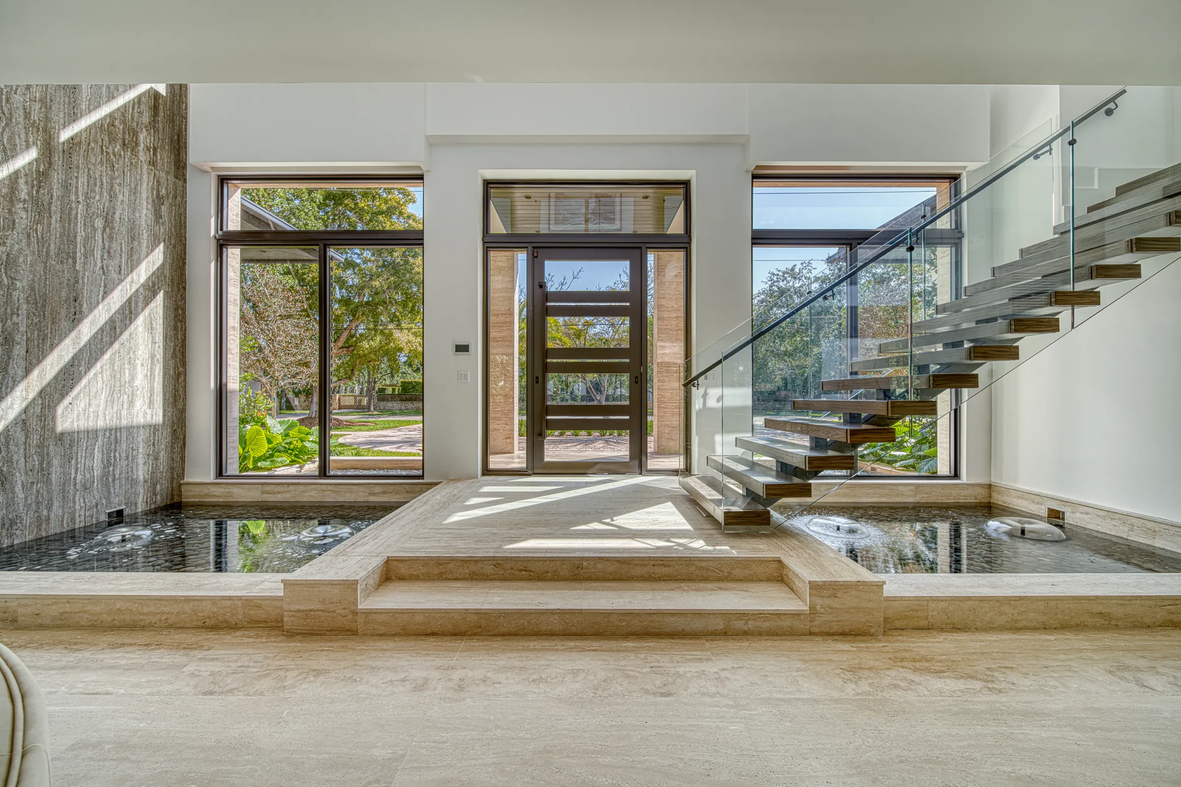 Sunlit travertine entry with floating glass staircase and reflecting pools